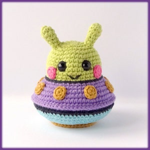 Puede incluir: Un extraterrestre verde de crochet con mejillas rosadas y ojos negros, sentado en un OVNI morado y azul con detalles marrones.