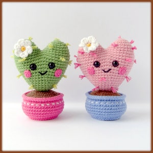 May include: Two crocheted heart-shaped cactus plants in pots. One cactus is green with a pink pot and the other is pink with a blue pot. Both have white flowers and smiling faces.
