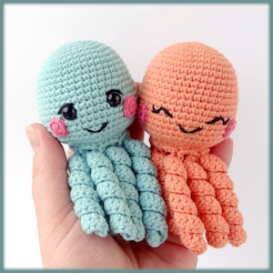 May include: Two crocheted octopus toys. One is light blue, the other is peach. Both have black eyes, pink cheeks, and smiling faces. The tentacles are curled. The toys are held in a hand.
