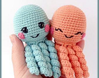May include: Two crocheted octopus toys. One is light blue, the other is peach. Both have black eyes, pink cheeks, and smiling faces. The tentacles are curled. The toys are held in a hand.