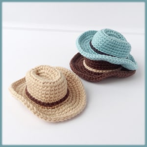 Pode incluir: Dois chapéus de caubói de crochê, um em bege com uma faixa marrom e outro em azul claro com uma faixa azul escura.