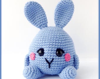 Könnte beinhalten: Ein gehäkelter blauer Hase mit rosa Wangen und schwarzen Augen. Der Hase sitzt auf einer weißen Oberfläche.