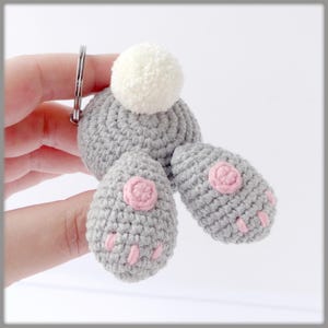 Puede incluir: Un llavero de conejito de crochet gris con detalles rosas y una cola de pompón blanco.