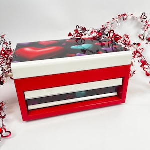 May include: Red jewellery box with a white lid and a drawer. The lid has a colourful print of hearts. The drawer has a clear front and shows colourful hearts inside.