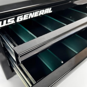 May include: Black metal tool chest with open drawers. The drawers have green dividers and are labelled "U.S. GENERAL" in white lettering. The open drawer reveals the interior of the tool chest.