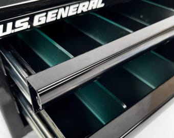 May include: Black metal tool chest with open drawers. The drawers have green dividers and are labeled "U.S. GENERAL" in white lettering. The open drawer reveals the interior of the tool chest.