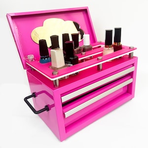 May include: A vibrant pink nail polish organiser with a handle and two drawers. The organiser has a cloud-shaped mirror and holds several bottles of nail polish. The organiser is made of wood and metal.