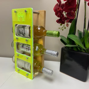May include: A neon green wine rack with four bottles of white wine. The rack has a wooden base and a clear glass front. The bottles are labeled with different names. The rack is sitting on a table with a plant in a black pot.