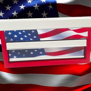 May include: A red-framed box with a white top and two white horizontal bars, featuring an American flag design. The stars and stripes are visible on the box. The background is a blurred image of the American flag.