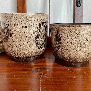May include: Two brown and white speckled ceramic plant pots. The larger pot is taller and wider than the smaller pot.