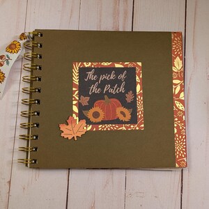 May include: A square spiral-bound scrapbook with a dark olive green cover. The center features a square label with the text "The pick of the patch" above a pumpkin and sunflower graphic. A decorative ribbon with sunflowers is tied to the gold wire coil.