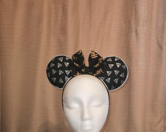 May include: Black and gold mouse ears with a bow and a Harry Potter themed fabric featuring the Deathly Hallows symbol and the words "Harry Potter".