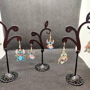 May include: Three pairs of dangle earrings featuring blue and pink cartoon characters with gold accents. The earrings are displayed on black metal stands.