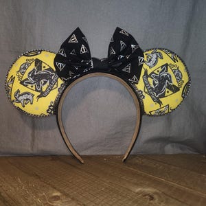 May include: A headband featuring two round yellow ears with a black Hufflepuff crest pattern and a central black bow decorated with silver Deathly Hallows symbols. The ears are edged with a thin, dark, braided trim, and the headband is a solid tan color.