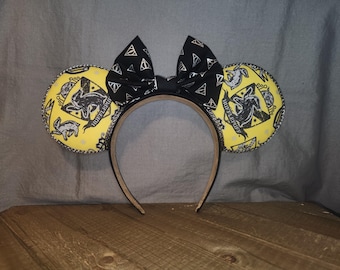 May include: A headband featuring two round yellow ears with a black Hufflepuff crest pattern and a central black bow decorated with silver Deathly Hallows symbols. The ears are edged with a thin, dark, braided trim, and the headband is a solid tan color.