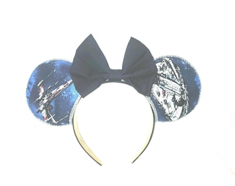 Puede incluir: Diadema de orejas de Minnie Mouse con un lazo azul marino. Las orejas presentan un diseño temático espacial con una nave espacial y un fondo estrellado. La diadema es de color dorado.