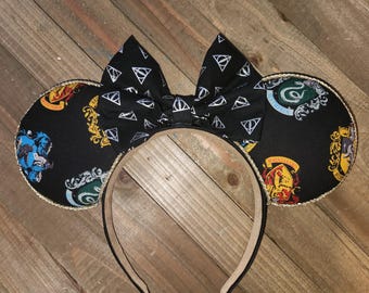 May include: Black mouse ears with a gold trim and a black bow with a white and silver pattern. The ears are decorated with the crests of the Hogwarts houses from the Harry Potter series.