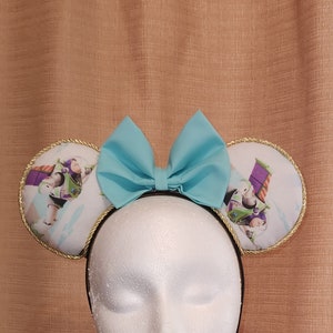 May include: A pair of light blue Minnie Mouse ears with a light blue bow. The ears are covered in a white fabric with a printed image of characters from the movie Toy Story. The headband is black.