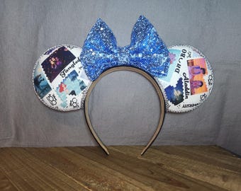 May include: A headband featuring two circular ears decorated with a white collage of Disney movie stamps, including Tangled and Aladdin, and a large, bright blue sequined bow centered at the top. The edges of the ears are lined with silver trim.