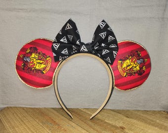 May include: A headband with two round red and gold ears featuring the Gryffindor crest and a black bow with white triangle symbols. The headband is black and beige.