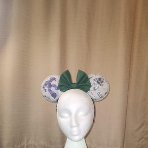 May include: Green fabric bow headband with white fabric ears featuring a cartoon character print. The ears are decorated with silver glitter.
