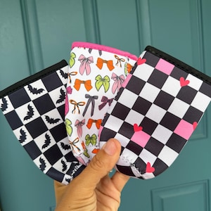 May include: Three beverage insulators with unique patterns are held up. One has a black and white checkerboard pattern with bats, another has bows in various colors, and the third has a black and white checkerboard pattern with pink hearts. Each has a black trim.