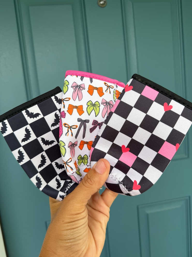 May include: Three beverage insulators with unique patterns are held up. One has a black and white checkerboard pattern with bats, another has bows in various colors, and the third has a black and white checkerboard pattern with pink hearts. Each has a black trim.
