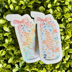 Pode incluir: Dois adesivos em forma de frasco com desenhos florais e la&ccedil;os cor-de-rosa. Um frasco tem glitter iridescente, o outro tem o nome "Alan" em it&aacute;lico. O fundo &eacute; um leito de folhas verdes.