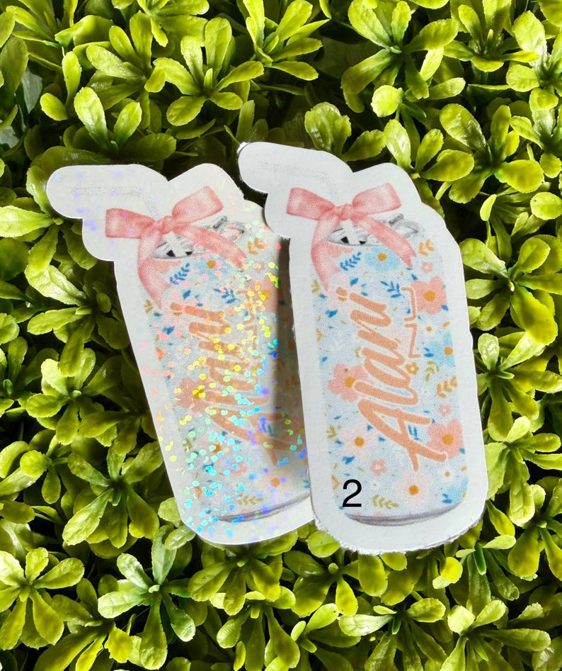 Pode incluir: Dois adesivos em forma de frasco com desenhos florais e la&ccedil;os cor-de-rosa. Um frasco tem glitter iridescente, o outro tem o nome "Alan" em it&aacute;lico. O fundo &eacute; um leito de folhas verdes.