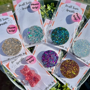 Peut inclure: Plusieurs supports de téléphone recouverts de paillettes dorées, turquoise, violettes et marron. Chaque support est emballé dans du plastique transparent avec une étiquette rose et blanche indiquant "Small Town Sparkle Phone Grip!"