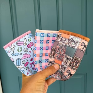 May include: Three fabric drink sleeves with different patterns are held up against a teal door. The patterns include a light blue with cowboy boots and cherries, a pink gingham, and a patchwork design with bows.