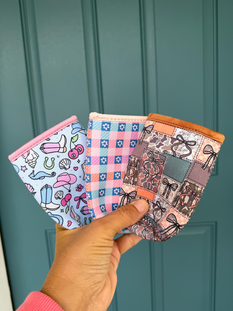 May include: Three fabric drink sleeves with different patterns are held up against a teal door. The patterns include a light blue with cowboy boots and cherries, a pink gingham, and a patchwork design with bows.