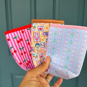 May include: Three colorful beverage insulators with unique patterns. One has pink stripes and cherries, another features postage stamp designs, and the third has a blue floral pattern. Each has a contrasting trim.