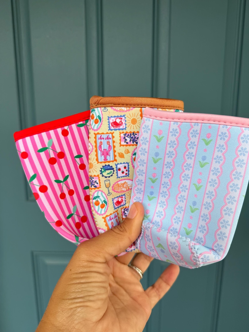 May include: Three colorful beverage insulators with unique patterns. One has pink stripes and cherries, another features postage stamp designs, and the third has a blue floral pattern. Each has a contrasting trim.