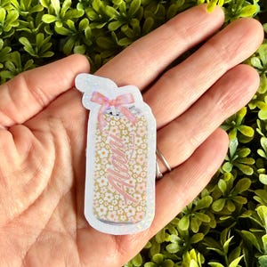Pode incluir: Um autocolante em forma de frasco com o nome "Alani" em escrita rosa. O frasco &eacute; preenchido com pequenas flores brancas sobre um fundo dourado e coberto com um la&ccedil;o rosa.