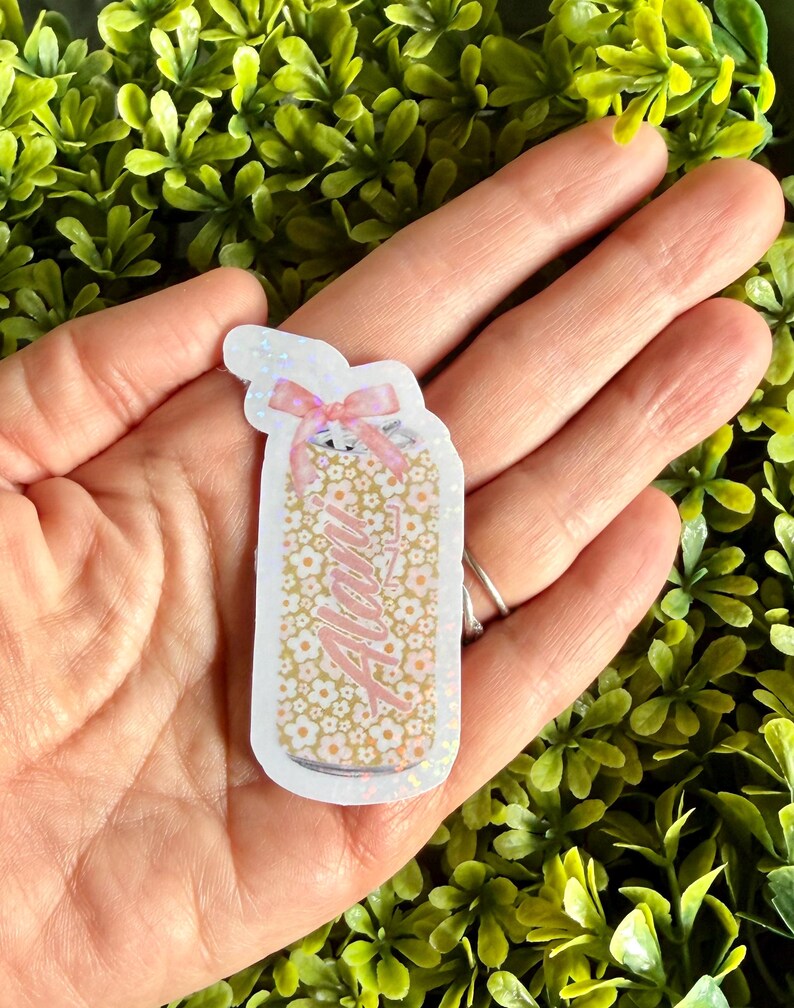 Pode incluir: Um autocolante em forma de frasco com o nome "Alani" em escrita rosa. O frasco &eacute; preenchido com pequenas flores brancas sobre um fundo dourado e coberto com um la&ccedil;o rosa.