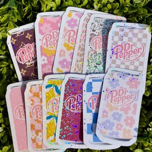 May include: A collection of Dr. Pepper can-shaped stickers with various designs. The stickers feature floral patterns, glitter, and checkered patterns in pink, yellow, blue, and brown. The Dr. Pepper logo is visible on each sticker.