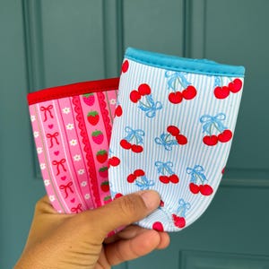 May include: Two fabric drink sleeves. One is pink with a strawberry and bow pattern, the other is blue and white striped with a cherry pattern. Both have a contrasting trim.