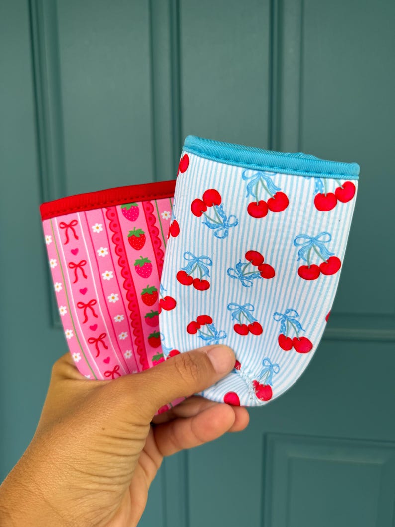 May include: Two fabric drink sleeves. One is pink with a strawberry and bow pattern, the other is blue and white striped with a cherry pattern. Both have a contrasting trim.