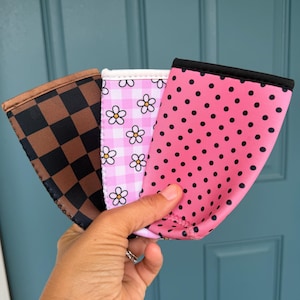 May include: Three beverage sleeves with different patterns are held up against a blue door. One sleeve has a brown and black checkered pattern, another has a pink and white floral pattern, and the last has a pink polka dot pattern.