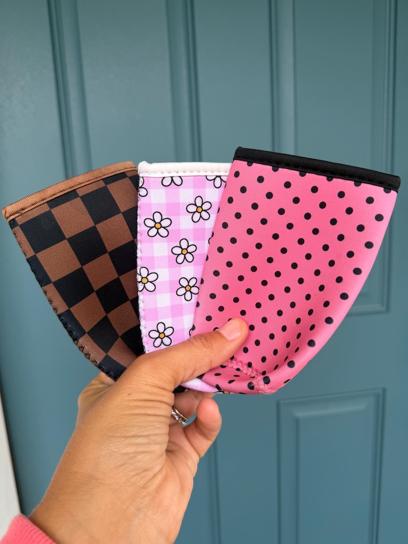 May include: Three beverage sleeves with different patterns are held up against a blue door. One sleeve has a brown and black checkered pattern, another has a pink and white floral pattern, and the last has a pink polka dot pattern.