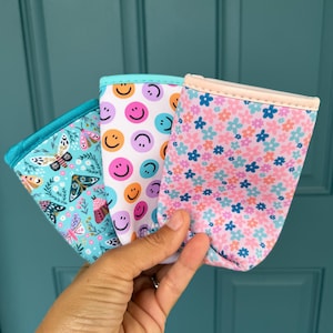 May include: Three colorful drink sleeves are held in front of a teal door. The sleeves feature different patterns: butterflies, smiley faces, and a floral design. Each sleeve has a contrasting trim.