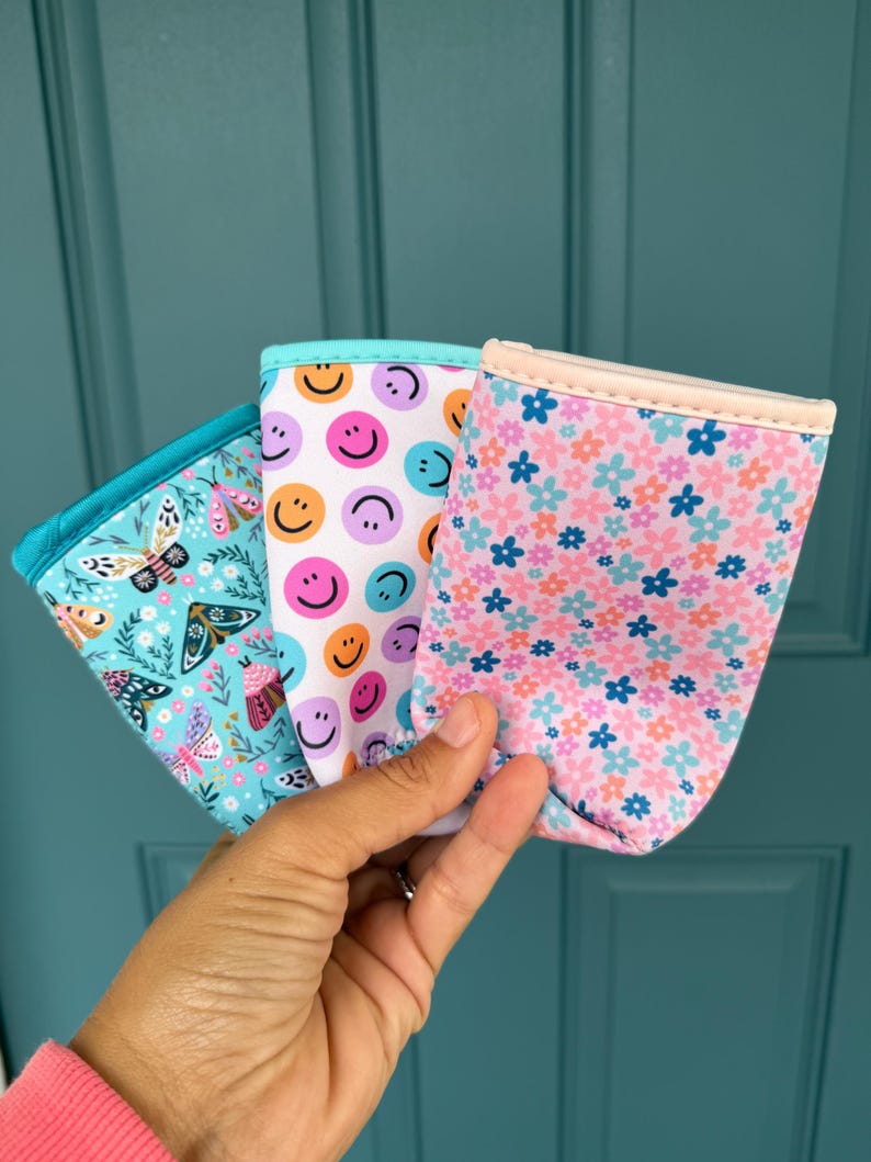 May include: Three colorful drink sleeves are held in front of a teal door. The sleeves feature different patterns: butterflies, smiley faces, and a floral design. Each sleeve has a contrasting trim.
