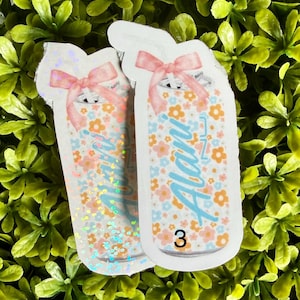 Pode incluir: Dois adesivos em forma de frascos, decorados com um padr&atilde;o floral em laranja, azul e rosa. Cada frasco tem um la&ccedil;o rosa no topo e o nome "Alani" em azul. O n&uacute;mero "3" est&aacute; na parte inferior.