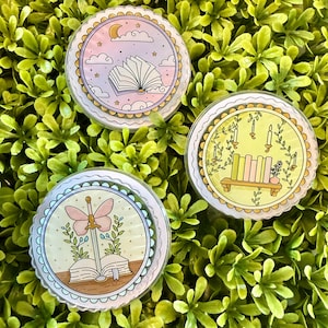 May include: Three round, clear stickers with whimsical illustrations. One features an open book with a moon and clouds, another shows candles and books, and the last depicts a sword with a butterfly and an open book. Each has a scalloped edge.