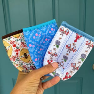 May include: Three fabric drink sleeves with different patterns are held up. One has a bee and floral design, another has a blue floral pattern, and the third has a patriotic theme with flags and lobsters. Each sleeve has a blue trim.