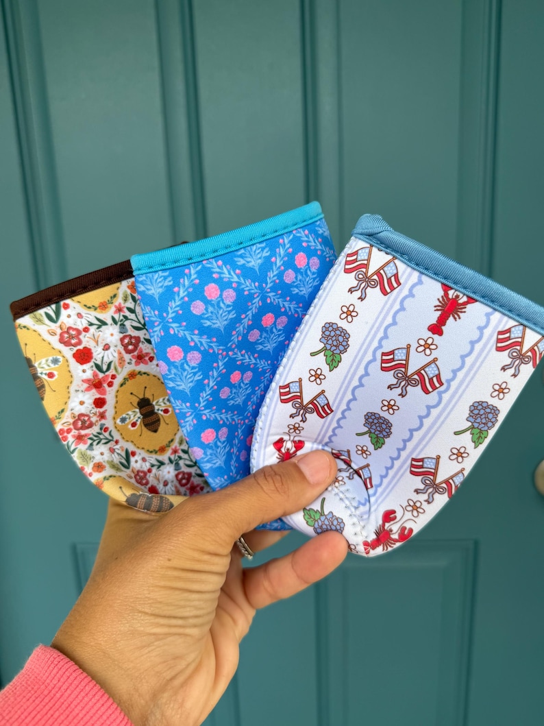 May include: Three fabric drink sleeves with different patterns are held up. One has a bee and floral design, another has a blue floral pattern, and the third has a patriotic theme with flags and lobsters. Each sleeve has a blue trim.