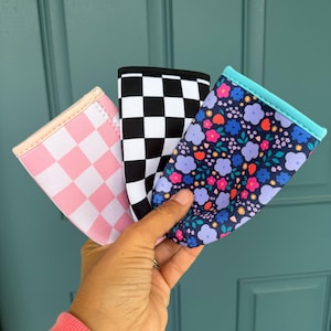 May include: Three beverage insulators with different patterns are held up against a teal door. One is pink checkered, one is black and white checkered, and the third has a floral pattern with blue, purple, and orange flowers.