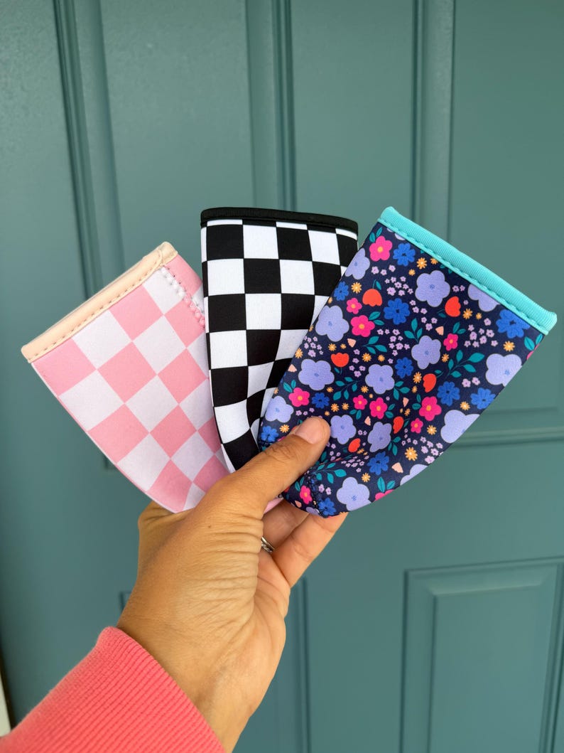 May include: Three beverage insulators with different patterns are held up against a teal door. One is pink checkered, one is black and white checkered, and the third has a floral pattern with blue, purple, and orange flowers.