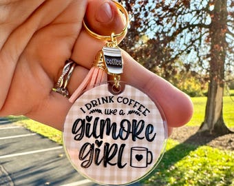 May include: A round keychain with the text "I drink coffee like a Gilmore Girl" in black script. The keychain has a gold ring, a pink tassel, and a coffee cup charm. The background is a tan and white plaid pattern.